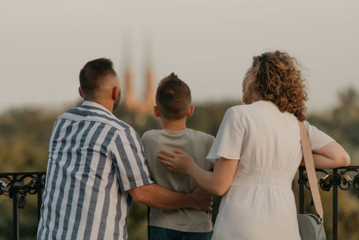 A photo from the back of father, mother, and son who are staring at the old European church. The family of tourists is enjoying the town sight in the evening. Tourists at sunset.