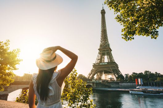 Young woman tourist in sun hat and white dress standing in front of Eiffel Tower in Paris at sunset. Travel in France, tourism concept. High quality photo