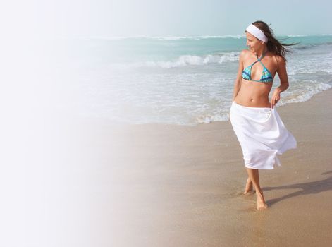 Beautiful young woman walks barefoot, on sandy beach, along ocean, wind is blowing her hair and white skirt. Concept of travel, relaxation, happiness and freedom, copy space