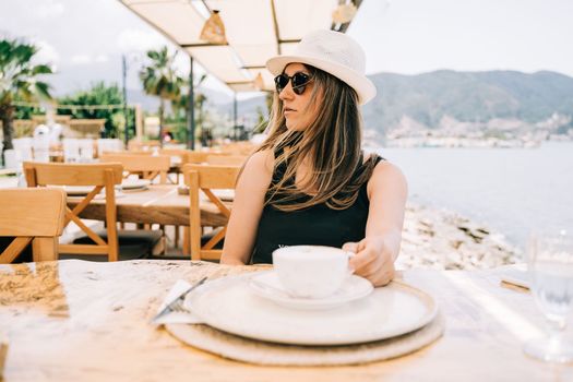 Young woman female in sunglasses and hat drinking coffee in a street outdoors seaside cafe restaurant coffee shop with scenery sea and mountains in the background. Hello summer holiday vacation.