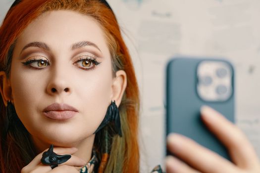 Woman takes selfie on front camera of smartphone. Female blogger runs a blog about fashion and makeup. Red-haired girl with Gothic-style jewelry with proud expression on her face. Witch aesthetic.