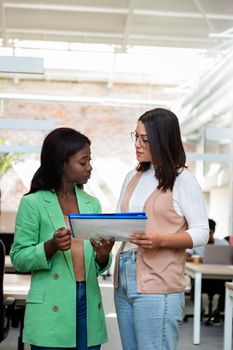 Two multiracial young women stand in the office to review some business documents. Copy space. Vertical image. Startup concept.