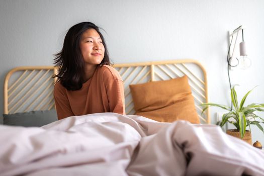 Happy young asian woman sitting on bed after waking up in the morning in beautiful, cozy bedroom at home. Lifestyle concept.