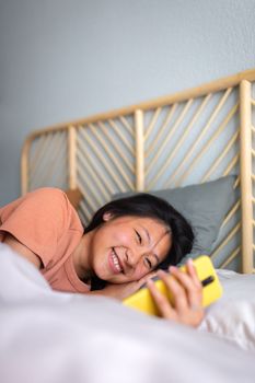 Happy teen girl using mobile phone in bed. Smiling Asian female looking mobile phone in the morning after waking up. Vertical. Technology and lifestyle.