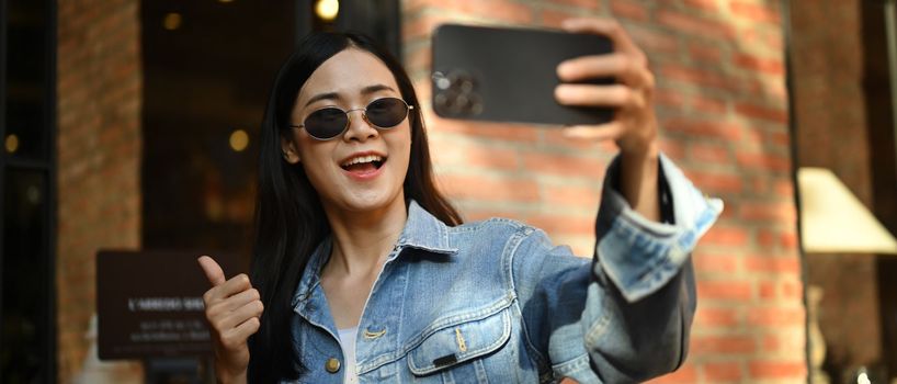 Cheerful stylish woman taking selfie or making video call on mobile phone while standing in city street.