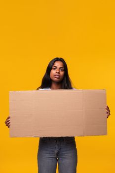 Indian woman with serious expression looking at camera holding a cardboard banner isolated in yellow background. Studio shot. Vertical image.