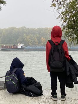 Thoughtful boys with hoods looking a cago ship sailing down Danube river. Cold and calm winter day concept