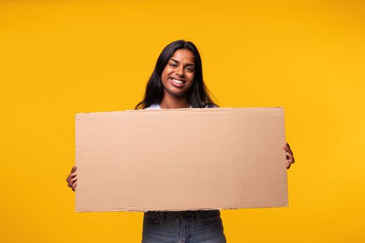 Young Indian asian woman looking at camera holding a cardboard banner isolated in yellow background. Studio shot. Activist concept.