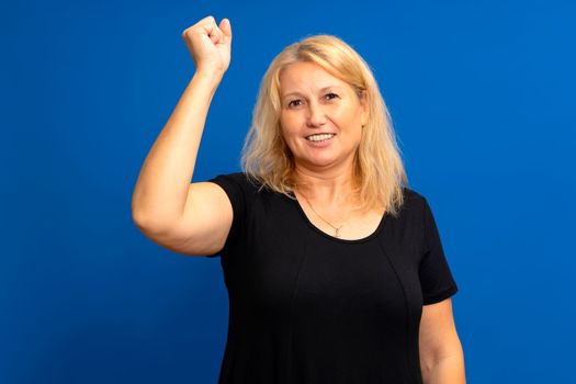 Excited Caucasian woman in her 40s with raised hand celebrating success. Portrait cheerful positive woman with raised fist. happy girl enjoy life blue color background