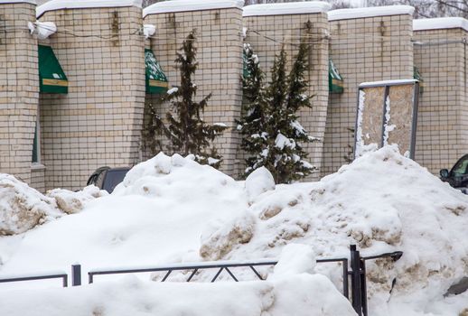 A large snowdrift against the background of a wall of a brick building. On the road lies white snow in high heaps. Urban winter landscape. Cloudy winter day, soft light.