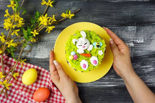 In the women's hands is a yellow plate with a beautifully decorated Easter cake. In the women's hands is a yellow plate with a beautifully decorated Easter cake, Easter decor. Easter bunny.