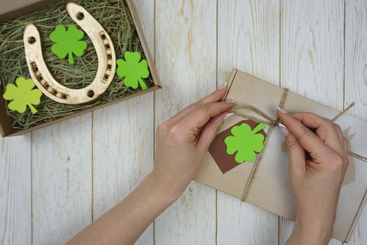 Women's hands are packing a gift for St. Patrick's Day. There is a golden horseshoe and clover leaves in the craft box.