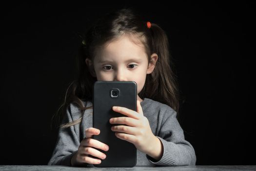 Problem of kids addiction to gadgets, online and screen time. Little girl looking at the screen of a big smartphone on black background.