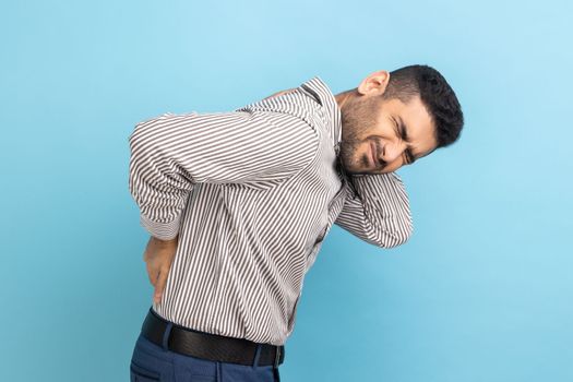 Backache or spine or kidney pain. Portrait of young sick businessman standing and touching his back and feeling pain, wearing striped shirt. Indoor studio shot isolated on blue background.