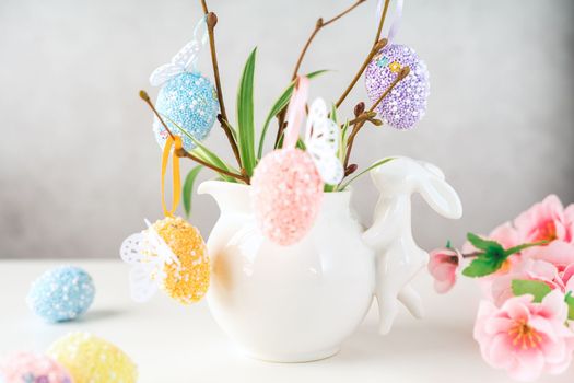 Home interior with easter decor. Vase with willow tree branches with Easter eggs and bunny on white table and background with copy space.