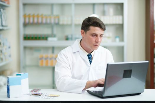 Male smart young-adult pharmacist in professional white gown working at desk in modern pharmacy, drugstore using laptop to check or record information. Medicine and healthcare concept.