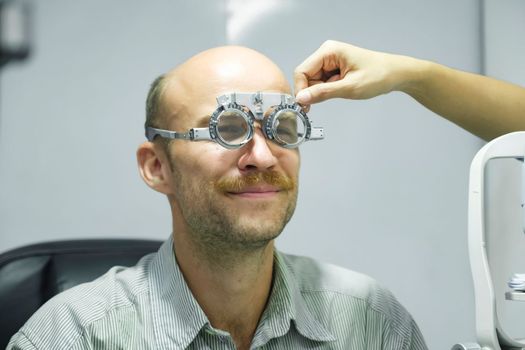 Smart handsome man sitting in optometrist cabinet having his eyesight checking, examining, testing with trial frame glasses by professional optician for new pairs of eyeglasses.