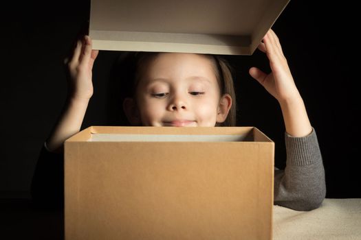 Emotional happy smiling little girl opens a long-awaited package with a gift, copy space on cardboard, look at box.