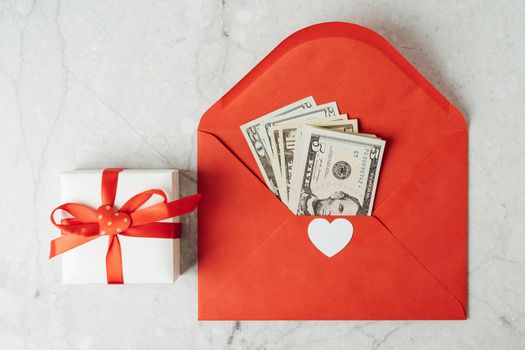 Red paper envelope with money dollars bills. Flat lay of gray working table background with Valentine gift, letter, heart shape. Top view, mock up greeting card. Valentine's Day budget
