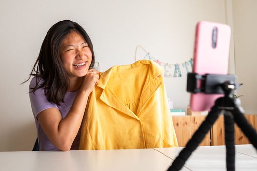 Happy Chinese young female fashion influencer showing clothing to followers during live stream using mobile phone on a tripod. Asian influencer recording video for social media.Video call.Technology.