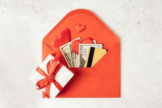 Red paper envelope with money dollars bills and credit card. Flat lay of gray working table background with Valentine gift, letter, heart shape. Top view, mock up. Valentine's Day budget
