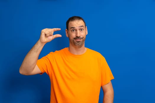 Hispanic man standing over blue background with skeptical face gesturing with hand making little sign with fingers looking and camera