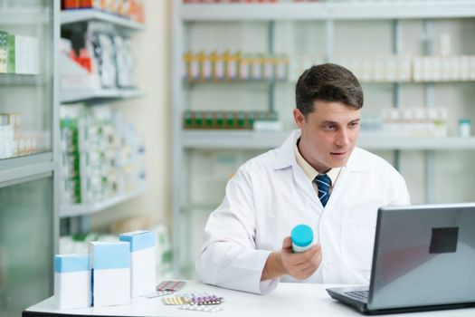 Male smart young-adult pharmacist in professional white gown working at desk in modern pharmacy, drugstore using laptop to check or record information. Medicine and healthcare concept.