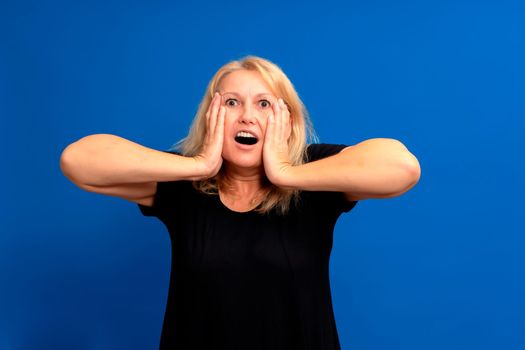 Caucasian blonde woman wearing a black t-shirt with her hands on her face excited and surprised isolated on blue studio background