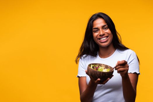 Happy young Indian woman eating healthy salad looking at camera. Isolated on yellow background. Studio shot. Healthy lifestyle concept.