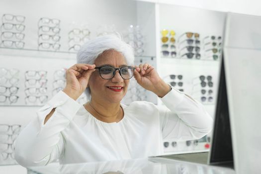 Woman trying, choosing, and deciding for the right frame for her new pair of eyeglasses looking at mirror after finishing with examinaiton in optic store.