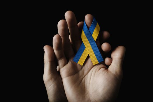 hand in hand holding a blue yellow ribbon on a dark background. concept needs help and support, truth will win