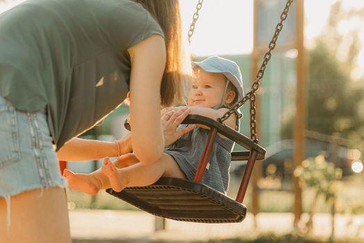 Toddler baby girl on a swing on the warm summer evening. Mother is swinging her young daughter on a sunny playground.