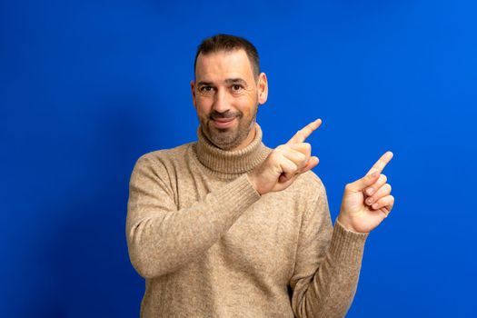 Portrait of a happy latin man in brown turtleneck pointing index fingers at copy space isolated over blue background