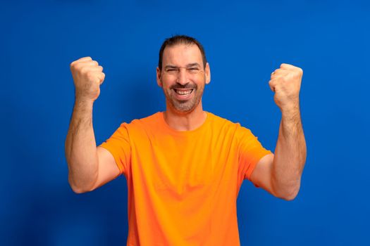 Excited joyful man in orange t-shirt standing isolated over blue background, celebrating success