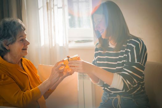 A young girl congratulates an elderly woman, her mother or grandmother, daughter holds in her hands a small cute beautifully wrapped gift for her grandparent, gives a surprise with love and care.