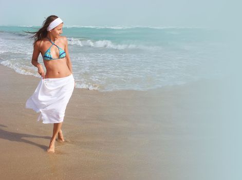 Beautiful happy girl walks barefoot, on sandy beach, along ocean, wind is blowing her hair and white skirt. Concept of travel, relaxation, happiness and freedom, copy space