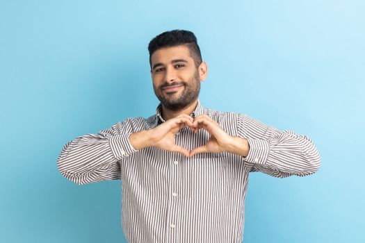 Happy positive handsome businessman with beard standing and showing heart shape making with hands, looking at camera with love, wearing striped shirt. Indoor studio shot isolated on blue background.