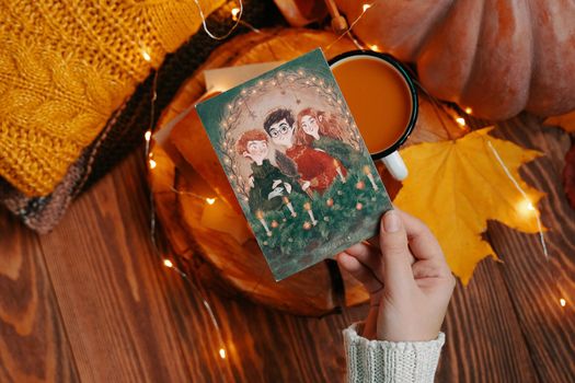 Postcard with illustration from Harry Potter in hand. Pumpkin, knitted sweater, autumn leaves and lights of garlands on a wooden background. Golden trio. Bishkek, Kyrgyzstan - November 1, 2019.