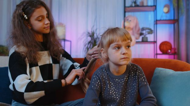Teenage and little sisters child kids using combs brushing make hairstyle coiffure. Female siblings children or best friends combing long hair at home play room. Family slumber party, plaiting hair