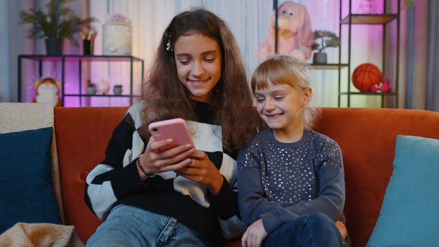 Teen sisters kids celebrate success win scream rejoices while doing online shopping on smartphone. Happy siblings children or best friends spend leisure time on social media app at home play room