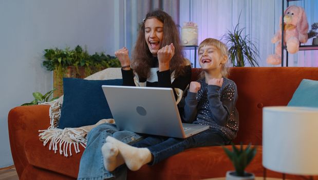 Overjoyed teenage and little sisters child kids playing on laptop raise hands in triumph winner gesture celebrate success win money in lottery game. Siblings children or best friends at home play room