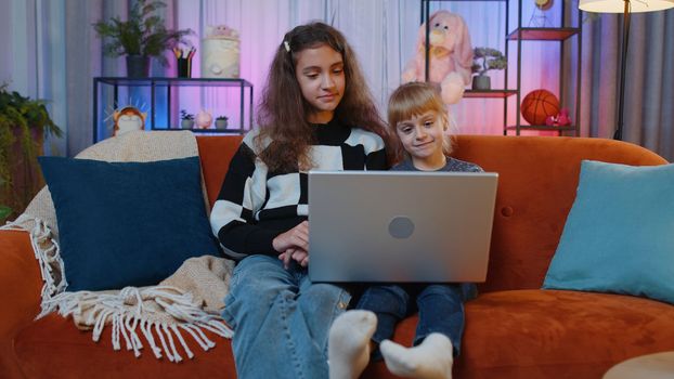 Teenage and little sisters child kids using laptop, making video webcam conference call, enjoying pleasant conversation. Female siblings children or best friends laughing, waving hello at home room