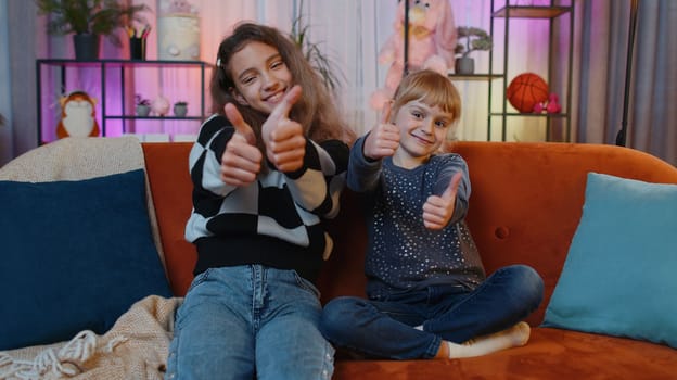 Like. Happy teenage child and little sister kid looking approvingly at camera showing thumbs up, like sign positive something good, positive feedback. Siblings children or friends at home play room