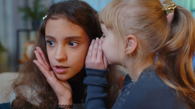Teenage child whisper news rumors in ear to little sister kid girl share secrets gossip having fun. Female siblings children or best friends at home play room. Friendship, family relationship concept