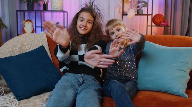 Child and little sister kid girls smiling friendly at camera, waving hands gesturing hello hi greeting or goodbye, welcoming with hospitable expression. Siblings children best friends at home playroom