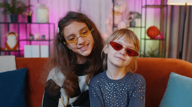 Portrait of happy smiling teenage child and little sister kid looking at camera wearing stylish sunglasses. Female siblings children or best friends sitting on sofa at home playroom. Friendship family