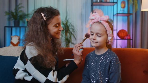 Teenage child helps or teaches to do face makeup her little sister kid girls. Female siblings children best friends putting applying make-up cosmetic at home play room. Friendship, family relationship