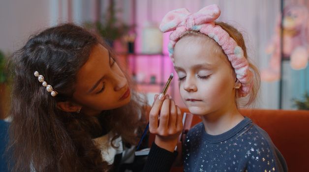 Teenage child helps or teaches to do face makeup her little sister kid girls. Female siblings children best friends putting applying make-up cosmetic at home play room. Friendship, family relationship