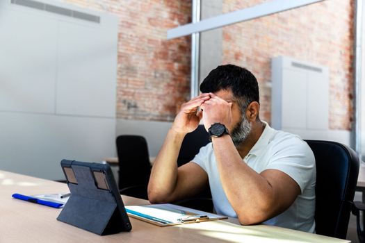 Mid adult Indian man worried and overwhelmed with work, forehead resting on hands at the office. Copy space. Depression at work.