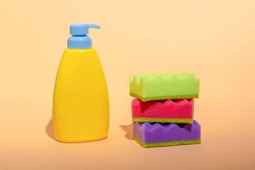 Stack of multi-colored dish wash sponges with bottle of soap on pastel orange background. Household cleaning scrub pad. Home cleaning concept